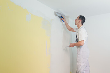 A working man applies putty to the walls in the apartment with a spatula, makes repairs in a new...