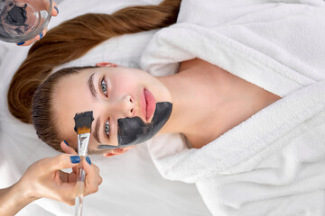 Obraz na płótnie Canvas people, beauty, spa, cosmetology and skincare. cute young woman at spa lying in white bathrobe, professional confident cosmetologist applying facial mask using brush. client look at camera top view