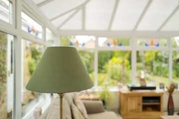 Shallow focus of lampshade seen in a large conservatory setting. Furniture is visible within the conservatory and in the rear a lush garden can be seen.