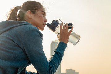 Fototapeta na wymiar Thirsty young woman runner is resting and drinking water bottle with headphone after morning cardio on street outdoors in the city background.
