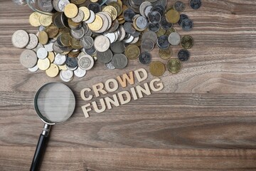 Crowd Funding text on the wooden table, with a magnifying glass and pile of coins.  