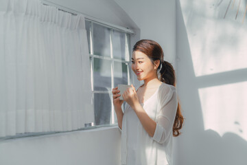 Young asian beautiful woman clothing white pyjamas and drink coffee beside window at morning in bedroom.