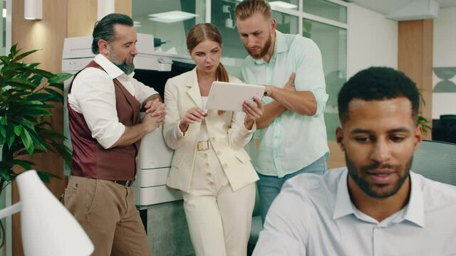 A beautiful blonde woman in a full white suit is talking to two of her workers in the office, while two other workers at their desks are doing their work and talking