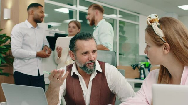 A kind looking old man is wearing a suit at the office and talking to a woman at the table about their work , in the back three workers are conversing near the printer. Arri Alexa Mini.
