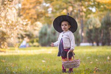 Little girl in witch costume at Halloween in autumn park with basket full of yellow leaves. Childhood, carnival.