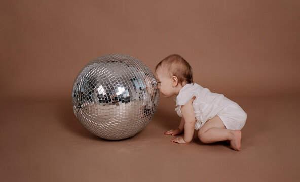 a baby girl in a white jumpsuit looks at a silver disco ball on a beige background with a place for text