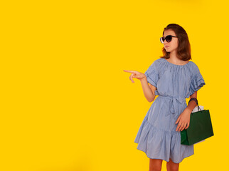 girl on a yellow isolated background with green and white packages in her hands in a blue dress and glasses shopping in a mall in a good mood and with an adorable smile. Place for your text. Advertisi