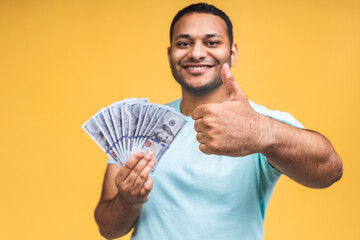 Winner! Young rich happy african american indian black man in casual holding money dollar bills with surprise isolated over yellow wall background.