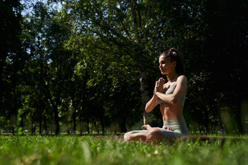 Naklejka na ściany i meble woman meditates in the park. a woman in a top and leggings in the summer in the park is engaged in yoga. pilates, gymnastics in the summer. sport exercises. young sports woman.