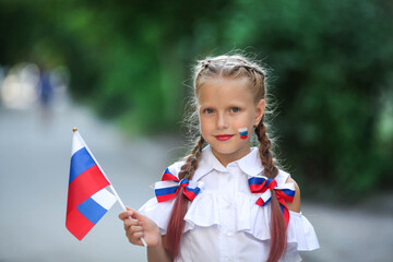 A school child stands on the outdoors and holds the flag of Russia. Little girl with face painting of Russian symbolism. Russian flag day.