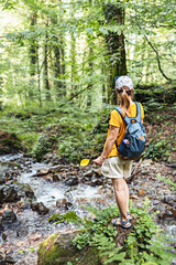 Rear view of young woman hiker in cap with backpack standing on mossy stone near mountain river in forest holding yellow fallen leaf in hand, walking in nature, eco tourism, hiking, outdoor activity