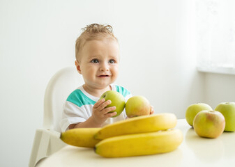 cute baby boy sitting at the table in child chair eating apple on white kitchen.