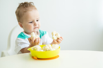 smiling baby boy sitting in a Childs chair eating cauliflower on white kitchen.