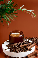 Opened brown cream jar on white gypsum cylinder podium display with palm leaves, coffee beans and cinnamon on pastel dark brown background for product presentation. Cosmetics beauty product promotion
