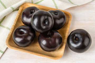Ripe sweet black plums on a square wooden plate over white wooden table. Large fresh whole plums close-up. Ready to eat juicy fruits and berries. Vegetarian food and healthy eating.