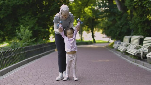 Positive Muslim mother and daughter with barbells in the summer park. Sporty fit Arab woman showing her little kid working out with dumbbells, making raises up