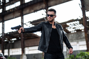 Man with a beard in a black leather jacket, wearing sunglasses and holding a gun. Man is aiming somewhere. Guns concept