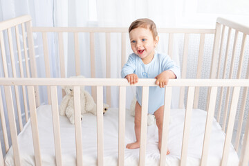 happy baby boy stands in the crib in the nursery and smiles or laughs