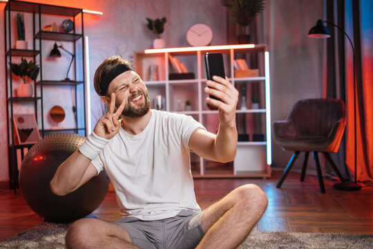Happy cheerful relaxed sporty bearded man sitting in lotus pose on the floor after home workout and making selfie of videochat with friend on smartphone, showing peace victory sign