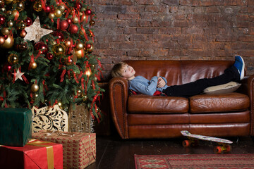 the boy is lying on the sofa near the Christmas tree . Happy boy waiting for Christmas presents . Cute baby boy at the christmas tree waiting for merry christmas . Xmas. Sale.