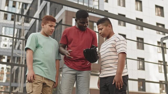 Low-angle medium shot of young African American man wearing baseball glove explaining how to catch ball to his two teenage sons listening to him, standing together at sports ground outdoors
