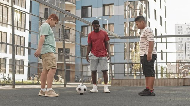 Full-length shot of young African American man and his two teenage sons standing in circle kicking football to each other during outdoor training at fenced sportsground in summer