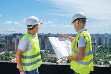 Two smiling mature men in a white helmet stand on the roof, hold a construction plan and showing...