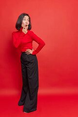 Surprised stylish dark haired Asian woman looks back with shocked expression wears turtleneck and loose black trousers poses against vivid red background copy space for your promotional content