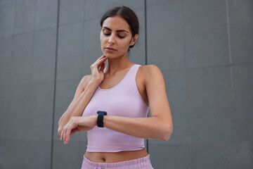 Serious dark haired European woman controls pulse after training looks at smartwatch wears cropped top poses against grey wall checks heart rate. Healthy lifestyle sport and workout concept.