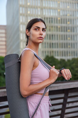 Pensive athletic Caucasian woman in sportsclothes checks time on smartwatch carries rolled karemat focused into distance plans her day stands on bridge admires cityscapes has training outdoor