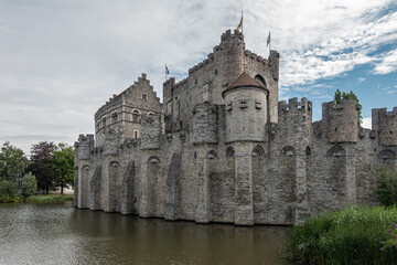 Fototapeta na wymiar Gent, Flanders, Belgium - July 30, 2021: West side of gray Gravensteem medieval stone castle under blue cloudscape behind dark water moat. Some green foliage.