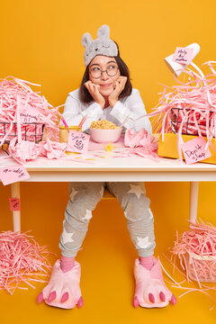 Dreamy Cute Asian Student Dressed In Soft Pajama And Sleepmask On Forehead Has Breakfast At Workplace Enjoys Cozy Home Environment Poses At Messy Desktop With Paper Rubbish Yellow Background