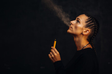 Obraz premium Beautiful young woman in black background with backlight smokes electronic cigarette