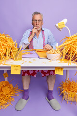 Shocked senior man work from home sits at white desk with folders paperclips bowl of cornflakes cut paper weras transparent glasses formal shirt tie around neck socks and slippers purple background