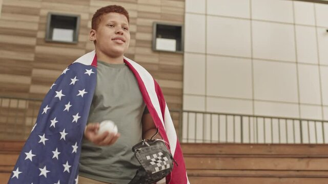 Low-angle medium shot of teenage red-haired boy wearing fabric US flag throwing baseball while talking to someone standing outdoors