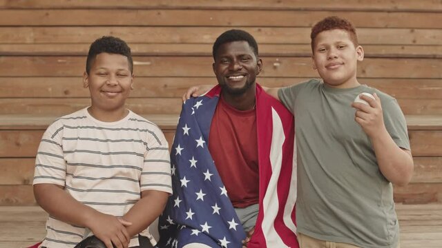 Medium portrait of smiling African American man smiling to camera with American flag on his shoulders while his two teenage sons on both sides posing with baseball and glove