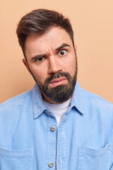 Obraz premium Close up shot of serious unshaven young European man looks attentively and puzzled raises eyebrows feels indignant reacts on something unexpected wears blue shirt isolated over beige background
