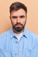 Obraz premium Vertical shot of handsome bearded man looks seriously has strict expression gazes directly at camera wears blue velvet shirt smirks face isolated over beige background. Human face expressions