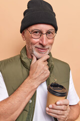 Pleased wrinkled man holds chin looks satisfied at camera has grey beard drinks takeaway coffee...