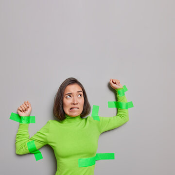 Worried Asian Woman Victim Of Harassment Stuck With Adhesive Plasters To Grey Wall Wears Green Turtleneck Bites Lips Has Nervous Expression Being Abused By Cruel Person. Copy Space For Your Text