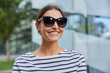 Carefree delighted young woman with dark hair chilld during nice spring day wears sunglasses striped jumper poses against blurred background focused away has upbeat mood broad smile white teeth