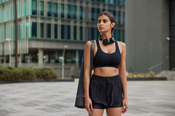 Horizontal shot of sporty woman in activewear carries rolled karemat prepares for training wears headphones around neck strolls at street against blurred background. Urban lifestyle concept.