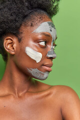 Profile shot of dark skinned woman with natural curly hair applies nourishing clay mask to refresh skin stands topless looks away poses against vivid green background. Face care and beauty concept