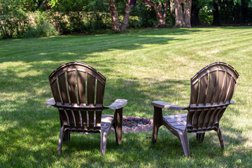 Close up backside view of a pair of gray lawn chairs in a back yard, near a fire pit, with light dappled shade