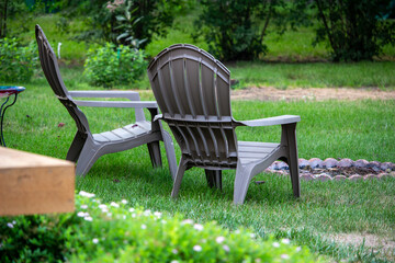 Close up backside view of a pair of gray lawn chairs in a back yard, near a fire pit, with light dappled shade