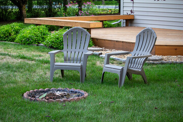 Close up view of a pair of gray lawn chairs in a back yard, near a fire pit, with light dappled...