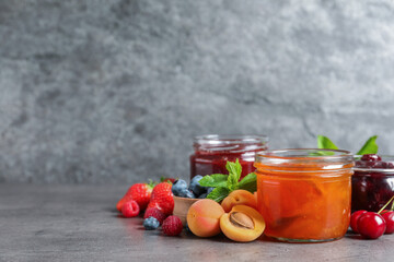 Jars with different jams and fresh fruits on grey table. Space for text