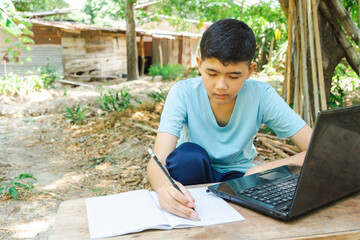 A boy is writing in a notebook while studying online with a laptop computer at home in the...