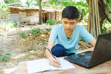 A boy is writing in a notebook while studying online with a laptop computer at home in the...