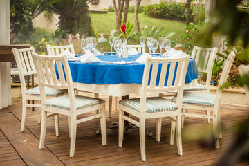 Outdoor restaurant table during the day with chairs, tableware, napkins and a red flower in the center - Corporate lunch or wedding party with garden in the back